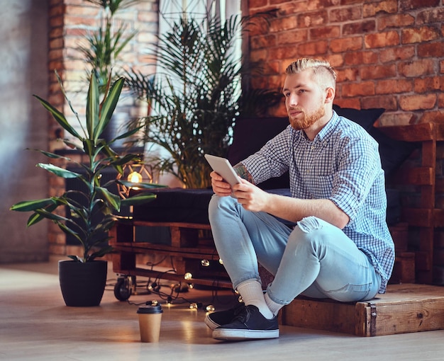 A imagem de corpo inteiro do homem elegante loiro vestido com uma camisa de lã e jeans usando um tablet PC em uma sala de estar.