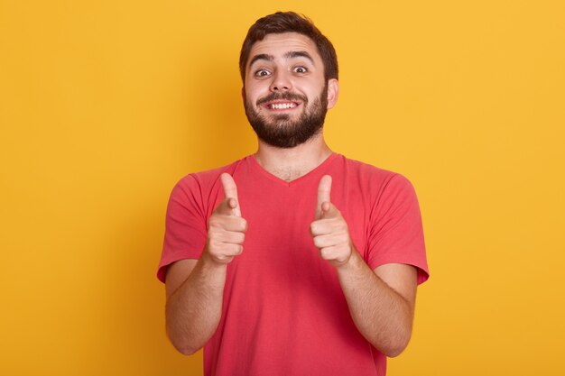 A imagem do homem moderno de sorriso considerável veste a camisa ocasional vermelha de t que mostra o sinal aprovado com ambos os polegares, levantamento do modelo isolado no amarelo, homem novo farpado com expressão facial feliz.