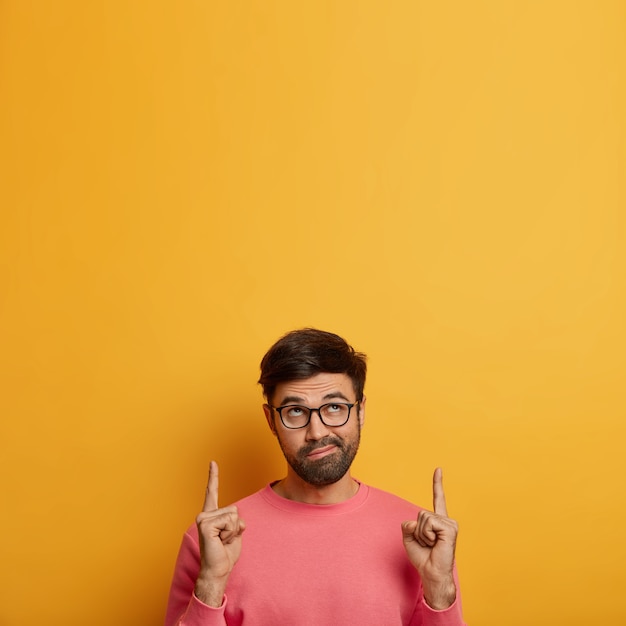 A imagem vertical de um homem barbudo intrigado aponta os dedos da frente para cima, franze os lábios e mostra algo interessante, usa um macacão rosado, isolado na parede amarela, mostra o produto. siga esta direção