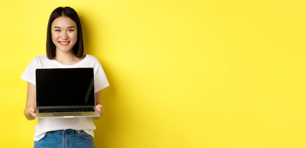 Foto grátis a jovem asiática demonstra a oferta online mostrando a tela do laptop e sorrindo em pé sobre ba amarelo