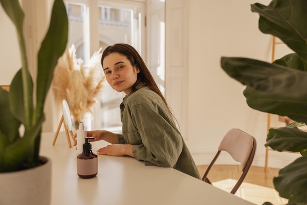 A jovem caucasiana moderna está sentada à mesa com um produto cosmético hidratante para cuidados com a pele morena de camisa verde olha para a câmera enquanto está no quarto conceito de cosméticos naturais