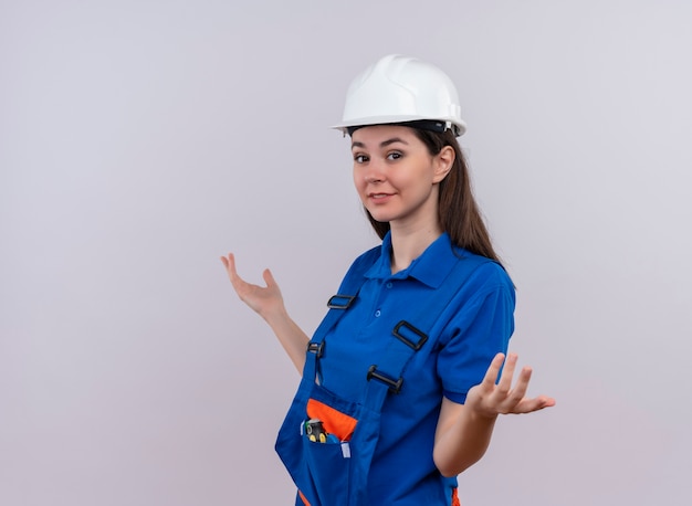 Foto grátis a jovem construtora confiante com capacete de segurança branco e uniforme azul fica de lado com as mãos para cima no fundo branco isolado com espaço de cópia