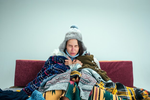 A jovem doente com gripe, sentado no sofá em casa ou no estúdio, coberto com roupas quentes de malha. Doença, gripe, conceito de dor. Relaxamento em casa. Conceitos de saúde.