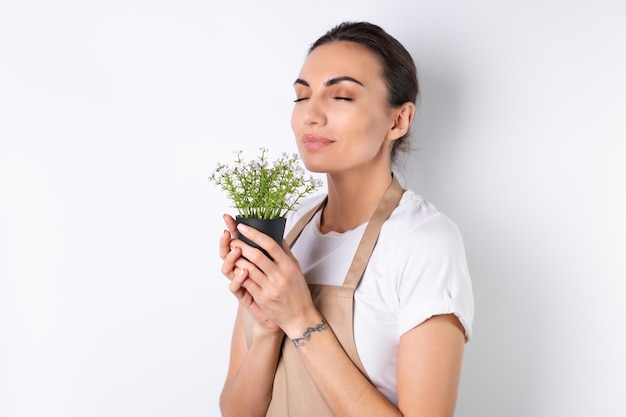 A jovem dona de casa em um avental em um fundo branco segura uma planta de casa em um pote sorri positivamente