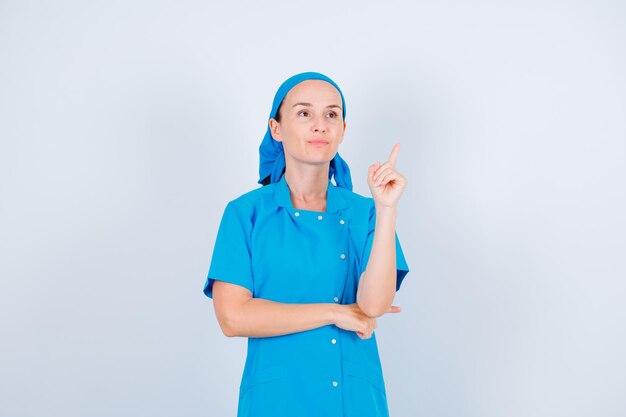A jovem enfermeira está olhando para cima e apontando para cima com o dedo indicador no fundo branco