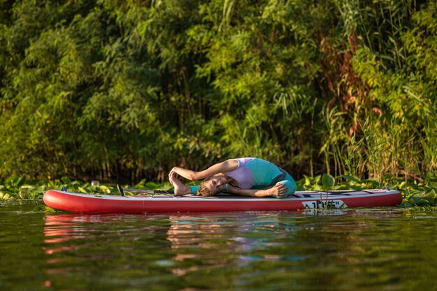 A jovem está fazendo ioga em uma prancha de stand up paddle SUP em um belo lago ou rio. O conceito de um estilo de vida saudável. Esporte. Ioga. Passatempo