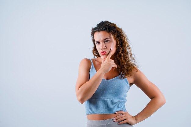 A jovem está pensando colocando a mão no queixo e colocando a outra mão na cintura no fundo branco