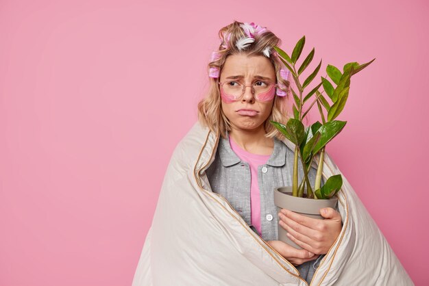 A jovem europeia triste e triste parece tristemente afastada aplica rolos de cabelo embrulhados em cobertor segura planta em vaso sente poses infelizes contra o espaço de cópia de fundo rosa para o seu conteúdo promocional
