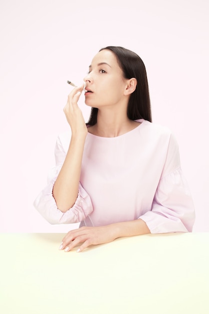 Foto grátis a jovem fumando cigarro enquanto está sentado à mesa no estúdio. cores da moda. o retrato de uma menina caucasiana em estilo minimalista com espaço de cópia