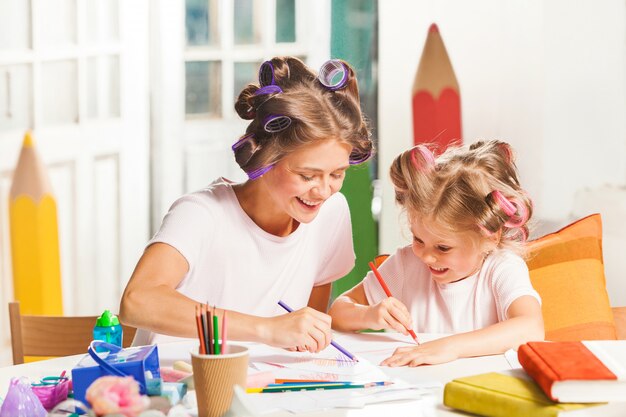 A jovem mãe e sua filha pequena desenhando com lápis em casa