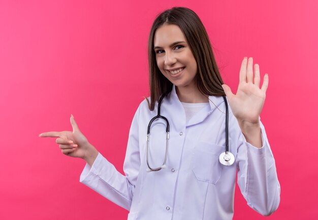 A jovem médica sorridente com um estetoscópio aponta para o lado e mostra um gesto de parada no fundo rosa isolado