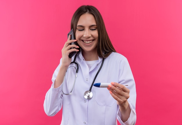 A jovem médica sorridente, usando um jaleco de estetoscópio, fala ao telefone, olhando para o termômetro com a mão no fundo rosa isolado