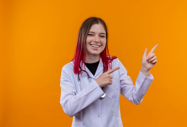 A jovem médica sorridente usando um manto médico de estetoscópio aponta o dedo para o lado em um fundo laranja isolado com espaço de cópia