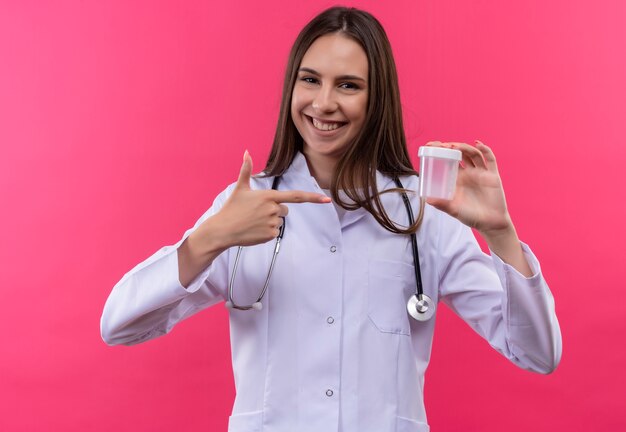 A jovem médica sorridente, usando uma bata médica de estetoscópio, aponta para uma lata vazia na mão, sobre fundo rosa isolado