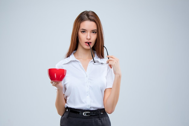 A jovem mulher de negócios de óculos com uma xícara de café vermelha sobre um fundo cinza