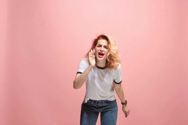 A jovem mulher irritada emocional gritando no fundo rosa studio