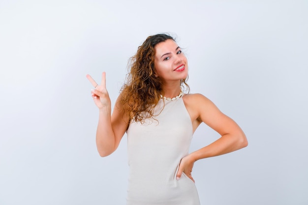 A jovem sorridente está mostrando dois gestos e colocando a outra mão na cintura em fundo branco