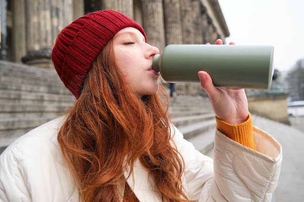 Foto grátis a jovem turista ruiva descansa durante sua viagem abre uma garrafa térmica e bebe chá quente fazendo uma pausa