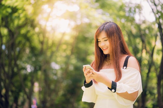 A linda mulher asiática está lendo mensagens de texto agradáveis ​​no telefone celular enquanto está sentado no parque no morno dia de primavera, linda mulher ouvindo música em fones de ouvido e pesquisando informações em celular.