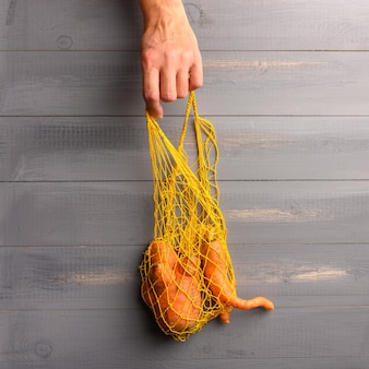 A mão de uma mulher pega um saco de barbante ecológico com uma cenoura feia na superfície de madeira