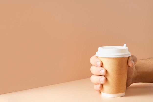 A mão do homem segura uma xícara de café com tampa fechada em um fundo cor de carne. copo de papel para bebida quente. ideia de maquete ou banner isolada na cor de fundo, espaço livre para texto
