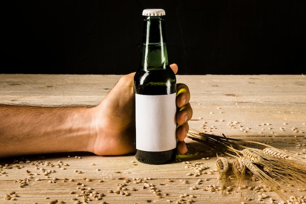 A mão do homem segurando a garrafa de cerveja com espigas de trigo na superfície de madeira