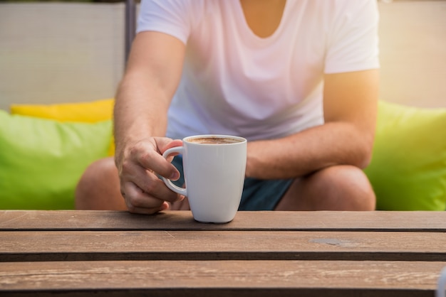 A mão está segurando uma xícara de café.Men estão bebendo café da manhã com um fundo verde lá fora. Man mãos segurando xícara de café no café ao ar livre Verão