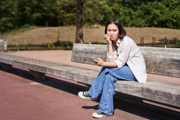 A menina asiática deprimida senta-se no banco no parque com o smartphone sentindo-se desconfortável e estressada franzindo a testa e