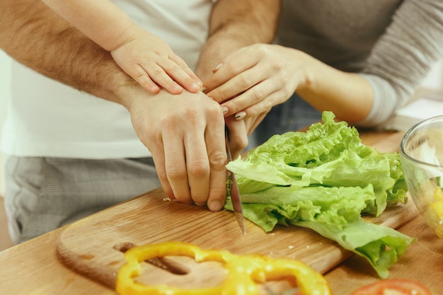 A menina bonitinha e seus lindos pais estão cortando vegetais e sorrindo enquanto fazem salada na cozinha de casa