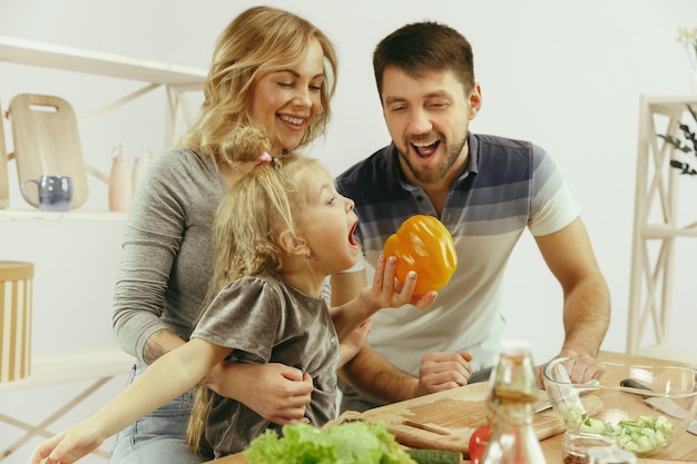 A menina bonitinha e seus lindos pais estão cortando vegetais e sorrindo enquanto fazem salada na cozinha de casa