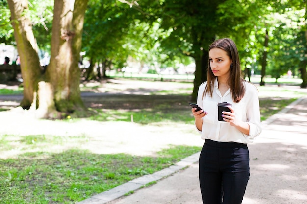 A menina caminha com o telefone na mão e uma xícara de café no parque