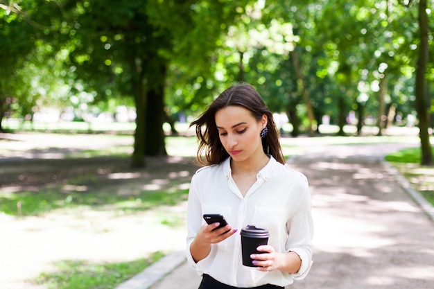 A menina caminha com o telefone na mão e uma xícara de café no parque