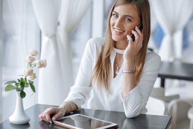 A menina de sorriso fala no telefone com um tablet na mesa