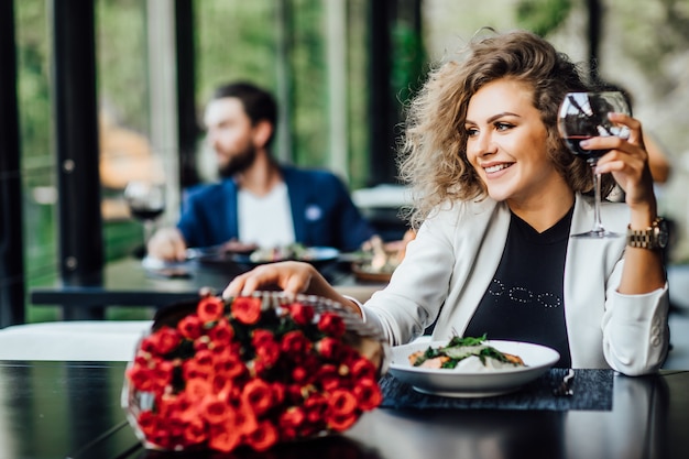 Foto grátis a menina está sentada a uma mesa no restaurante e bebe vinho, aprecia o perfume da flor de rosas e espera um encontro