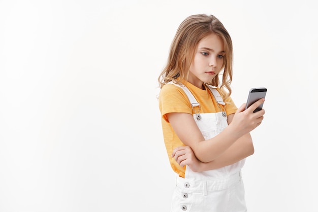 Foto grátis a menina loira bonita moderna usa um macacão de verão, segura o smartphone, fica relaxada com a tela do celular, rolar a internet