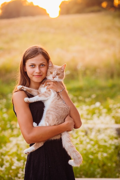 Foto grátis a menina no vestido preto segura um gato vermelho de pé no campo