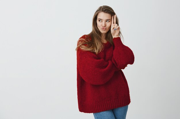 A menina quer chamar namoradas para ir festejar. Retrato de mulher feminina atraente na camisola de inverno elegante, dobrar os lábios no beijo, segurando o sinal v perto do rosto enquanto olha de lado