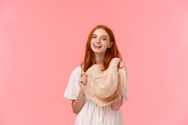 A menina ruiva macia, carismática e atraente, balançada na cintura, arrumada para as férias de verão, pronta para viajar para o exterior, segurando um chapéu de palha, rindo e sorrindo a câmera. fundo rosa de pé