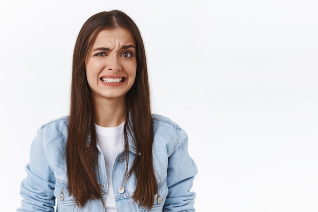 A menina se encolhe ao ver algo constrangedor e incomodativo. mulher fazendo sorriso desconfortável e semicerrando os olhos sentindo-se preocupada e descontente, ver situação incomodativa, fundo branco em pé