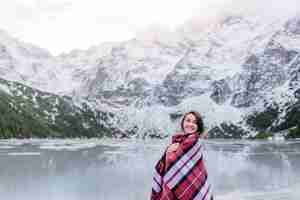 Foto grátis a menina sorrida coberta no cobertor quente está de pé perto do lago nas montanhas, jovem viajante