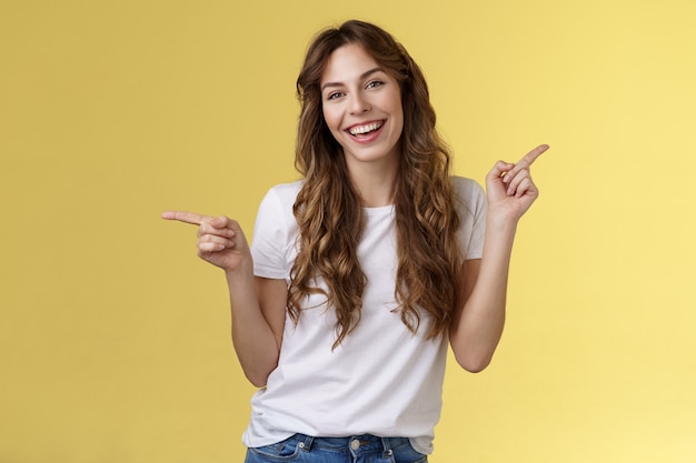 Foto grátis a menina tem duas sugestões apontando para os lados. mulher atraente de cabelos cacheados carismática e alegre apontando o dedo indicador esquerdo e direito apresentar produtos promocionais com um anúncio de recomendação com um sorriso amplo e animado.