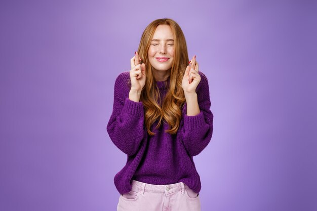 A menina transforma o desejo em realidade. Uma jovem ruiva atraente, esperançosa e positiva, com um suéter roxo, fecha os olhos e sorri como dedos cruzados para dar boa sorte, rezando e antecipando o milagre.