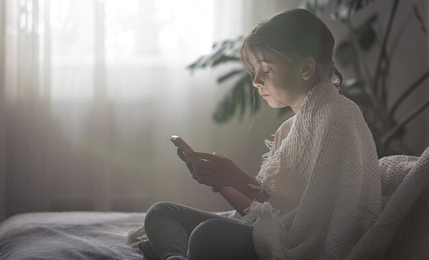 A menina usa o telefone enquanto está sentada na cama à noite, copie o espaço