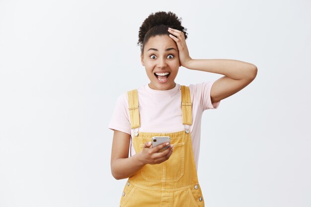 A menina verificou o calendário, lembrou-se do grande evento. Retrato de uma linda mulher afro-americana impressionada e surpresa de macacão amarelo, tocando o cabelo e sorrindo de empolgação, segurando o celular