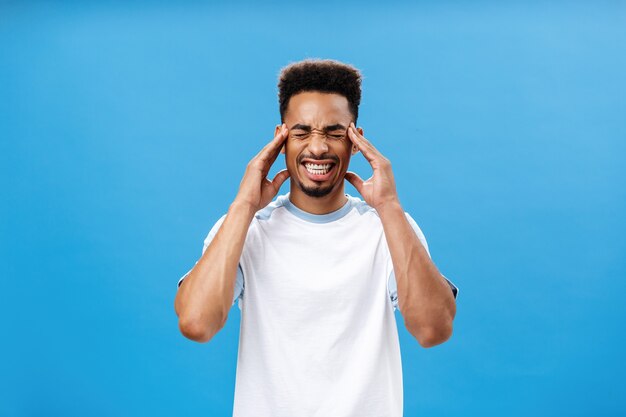 A mente enlouquece com pensamentos irritantes. Retrato de homem barbudo afro-americano bonito e intenso com corte de cabelo afro, cerrando os dentes e fechando os olhos, segurando as têmporas, sofrendo de mirgaine ou dor de cabeça