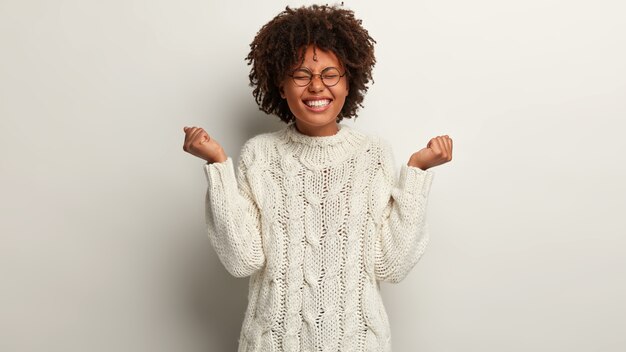 A metade da foto de uma mulher otimista se sente vencedora, levanta as mãos em punhos, vestida com um suéter de malha branco, fecha os olhos de prazer isolado sobre a parede branca. Pessoas, sucesso, conceito de alegria