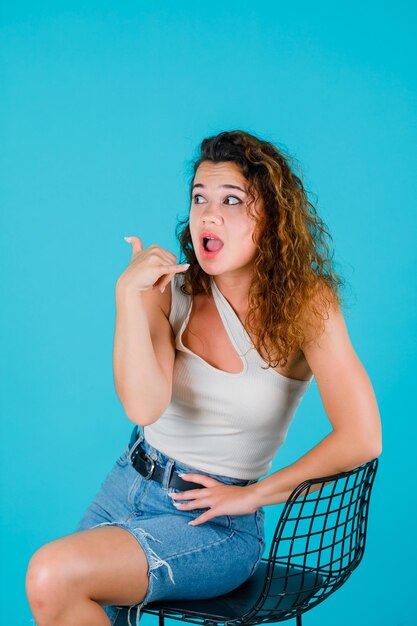 A moça está mostrando o gesto do telefone sentando-se na cadeira no fundo azul