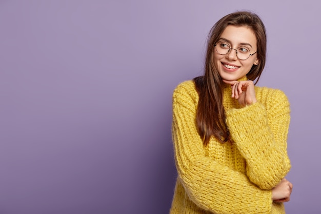 A modelo feminina caucasiana sorridente toca suavemente o queixo, olhando de lado com um sorriso cheio de dentes