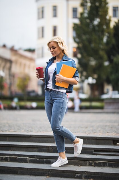 A modelo loira está indo para as aulas de trabalho pelo centro da cidade segurando notebooks e computador de café nas mãos pela manhã