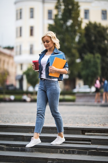 A modelo loira sorridente está indo para as aulas de trabalho pelo centro da cidade segurando notebooks, computador e café nas mãos pela manhã